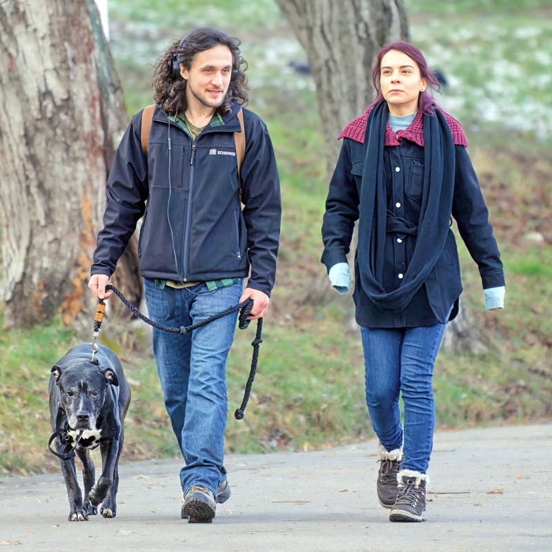 12 - Young couple walking a black dog on a paved path outdoors